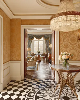 Elegant hallway with gold damask wallpaper, a round marble-topped table holding a vase of flowers, a crystal chandelier, and a view into a sitting room with blue drapes and classic furnishings.