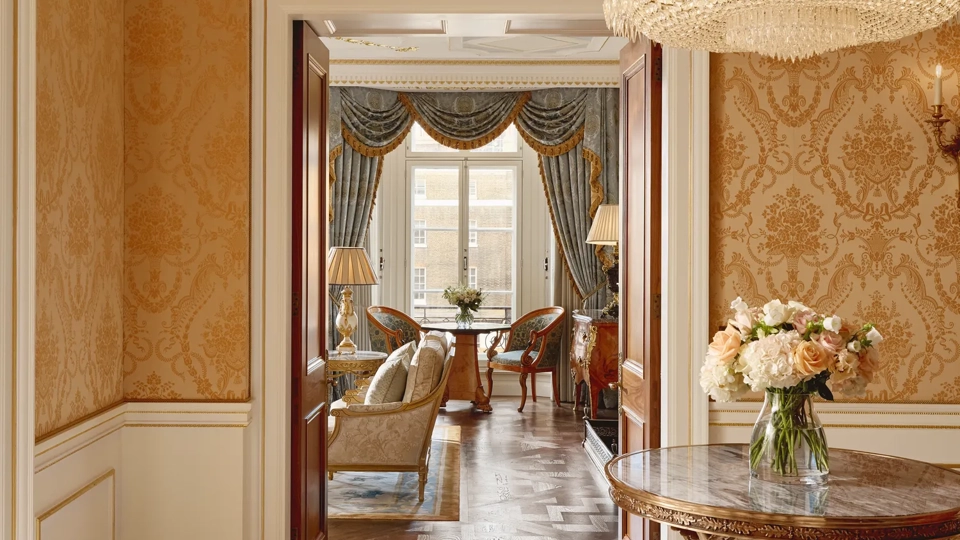 Elegant hallway with gold damask wallpaper, a round marble-topped table holding a vase of flowers, a crystal chandelier, and a view into a sitting room with blue drapes and classic furnishings.