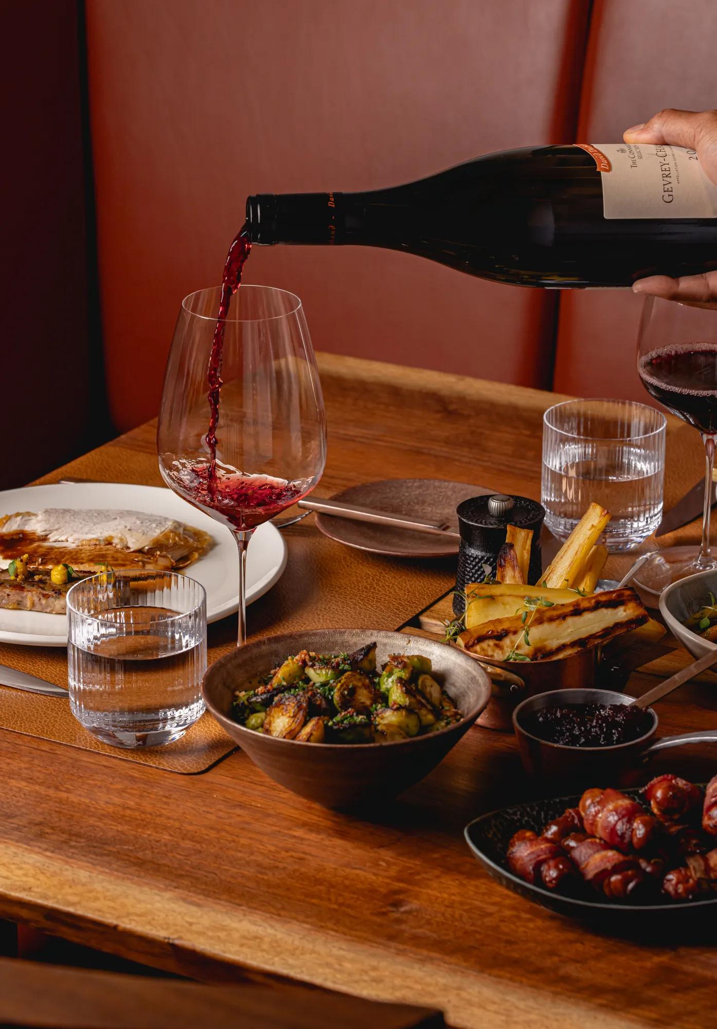Red wine being poured into a glass beside plates of roasted vegetables, sausages, and a main course on a wooden dining table.
