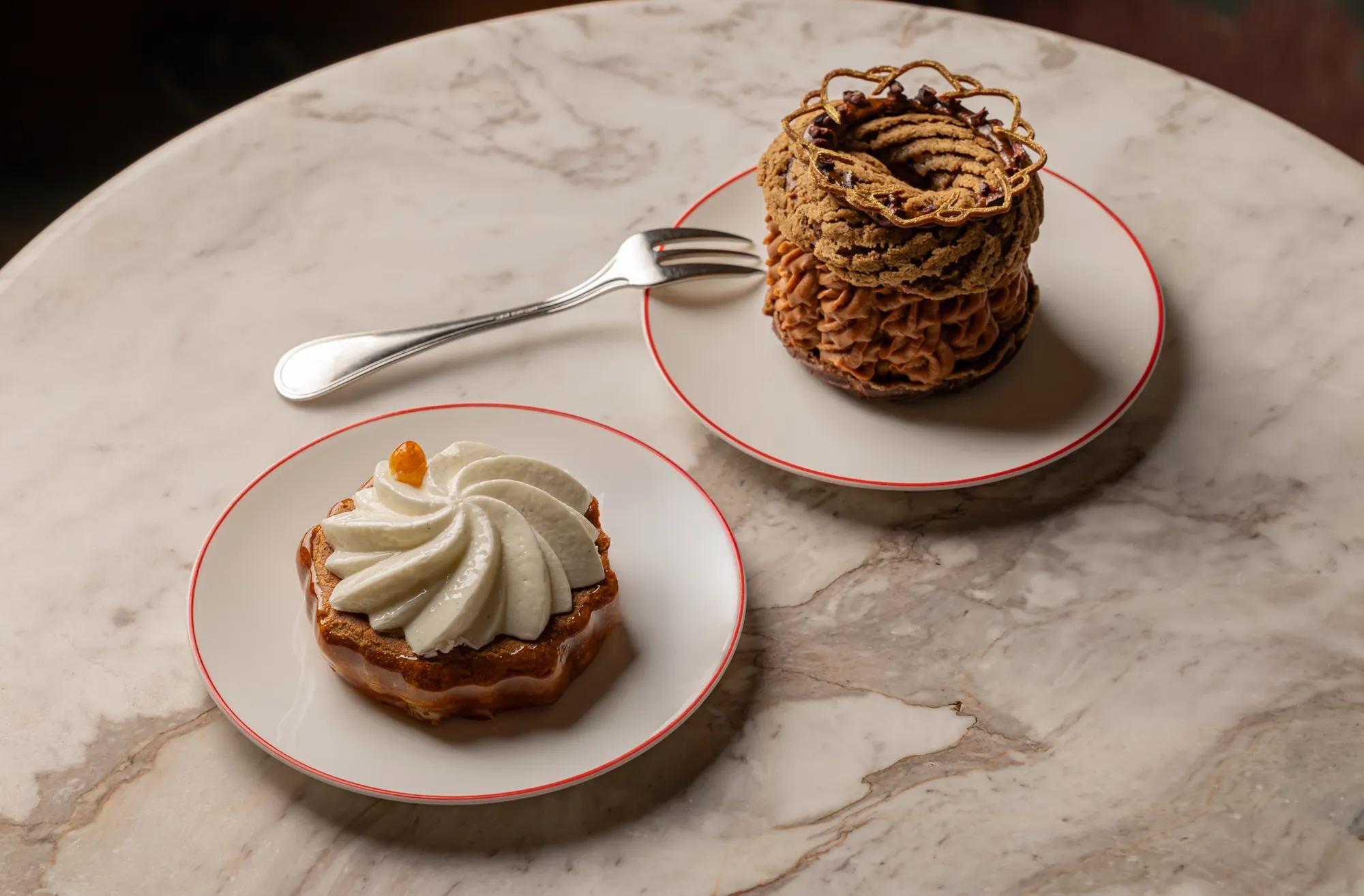 Two plated pastries on a marble table: a cream-topped tart and a chocolate Paris-Brest, with a fork placed between them.