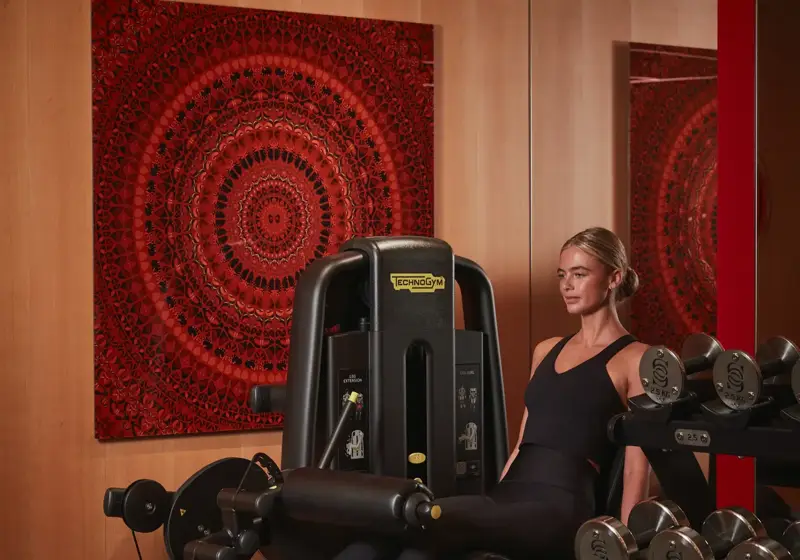 Person using leg extension machine in gym, with dumbbell rack and artwork behind.