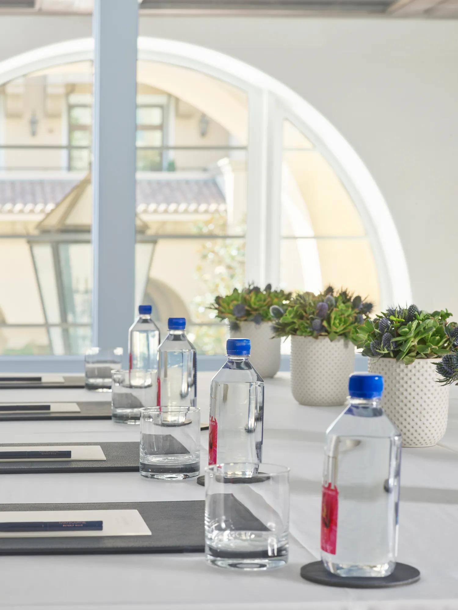 Conference table setup in Wilshire breakout rooms with water bottles, tumblers, notepads, and potted succulents beside a large arched window.