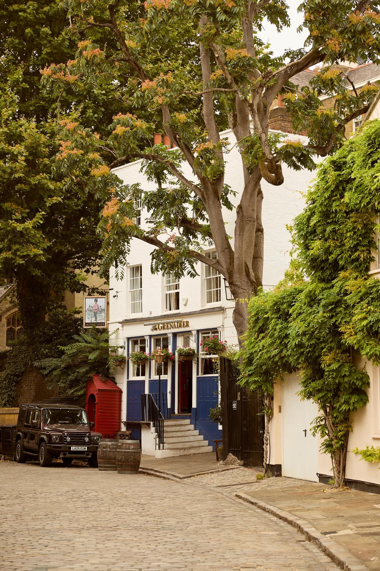Quiet cobbled street lined with trees, a white pub building, and a parked vehicle in a leafy residential setting.
