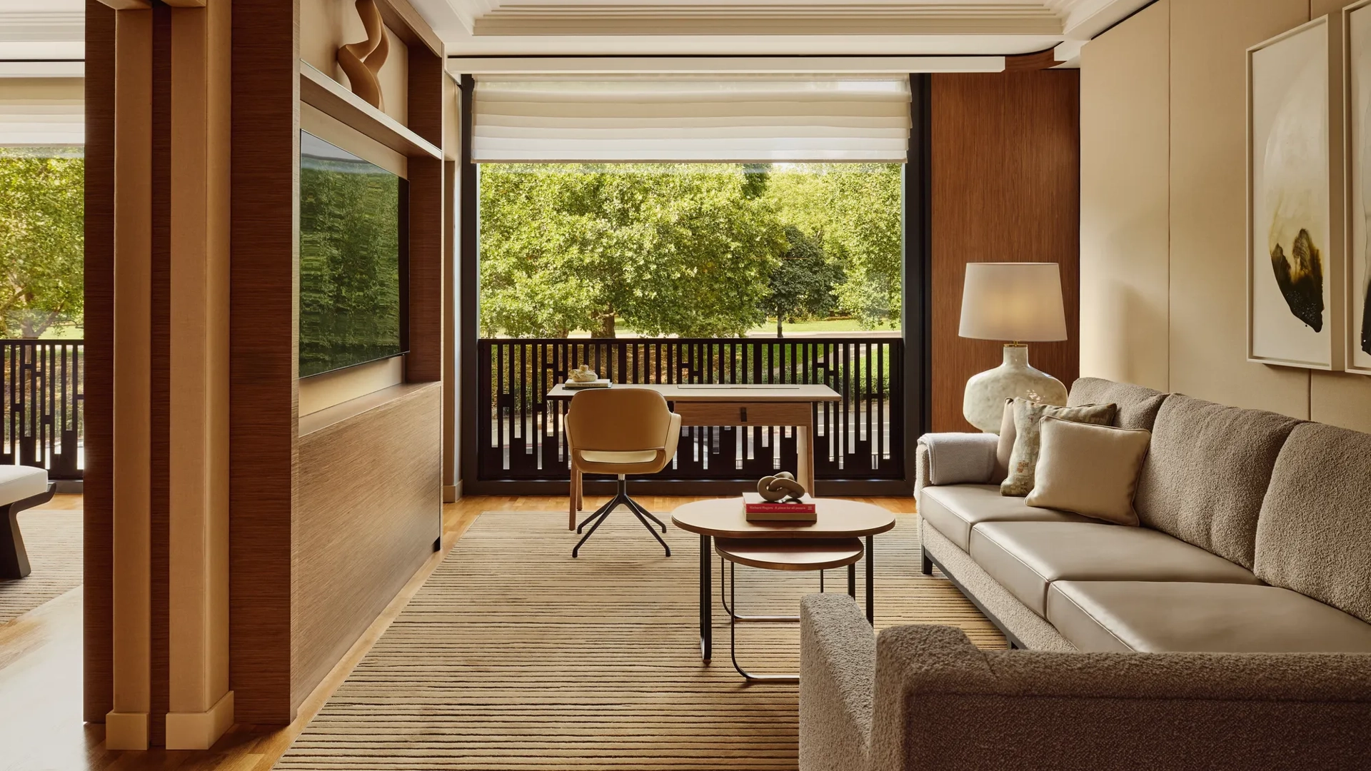 Living room with a beige sofa, desk facing a balcony, and large windows overlooking leafy park views.