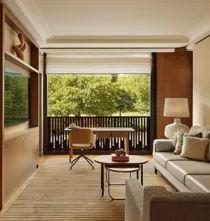 Living room with a beige sofa, desk facing a balcony, and large windows overlooking leafy park views.