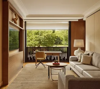 Living room with a beige sofa, desk facing a balcony, and large windows overlooking leafy park views.