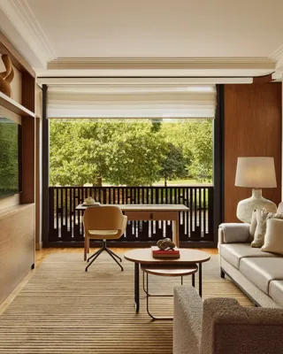 Living room with a beige sofa, desk facing a balcony, and large windows overlooking leafy park views.