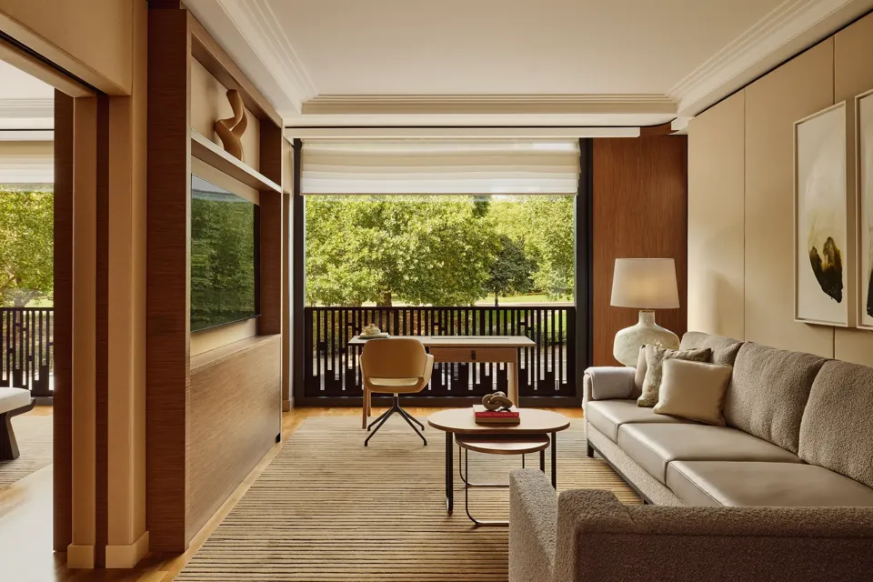 Living room with a beige sofa, desk facing a balcony, and large windows overlooking leafy park views.