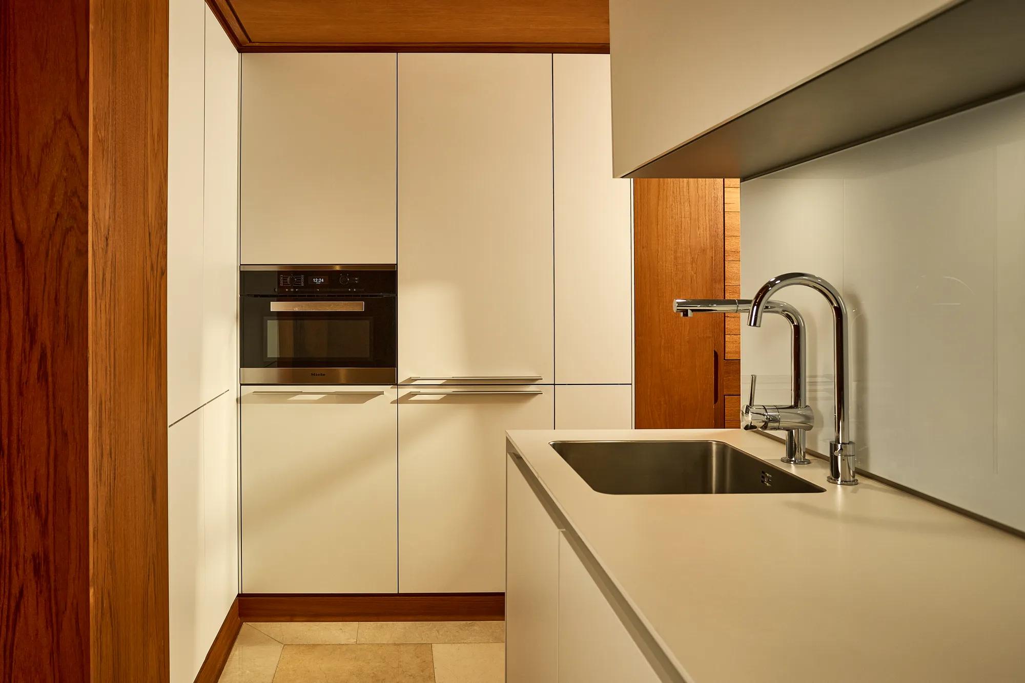 Compact kitchenette with white cabinetry, integrated oven, and sink with chrome tap beneath warm lighting.
