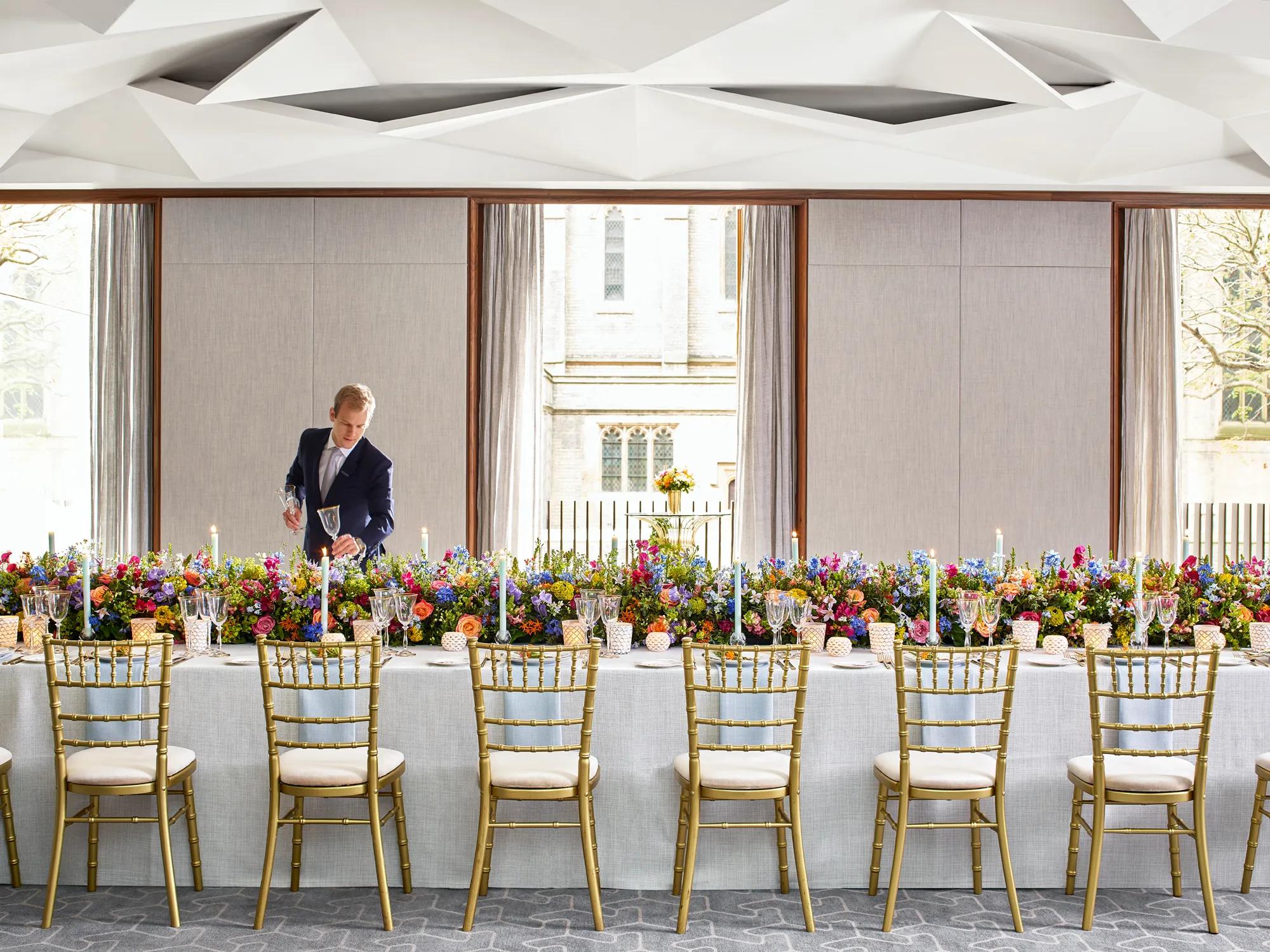 Long banquet table set with gold chairs, colourful flowers, and candles, with a server adjusting glassware