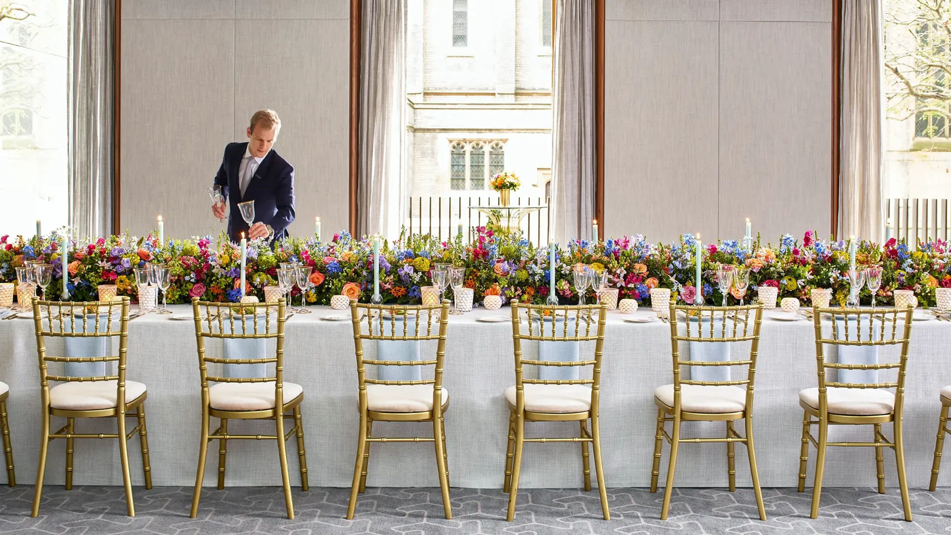 Long banquet table set with gold chairs, colourful flowers, and candles, with a server adjusting glassware