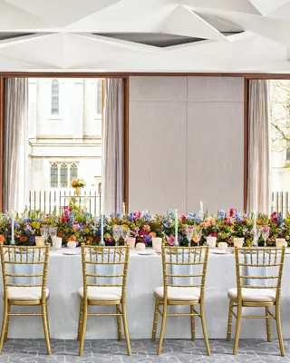 Long banquet table set with gold chairs, colourful flowers, and candles, with a server adjusting glassware