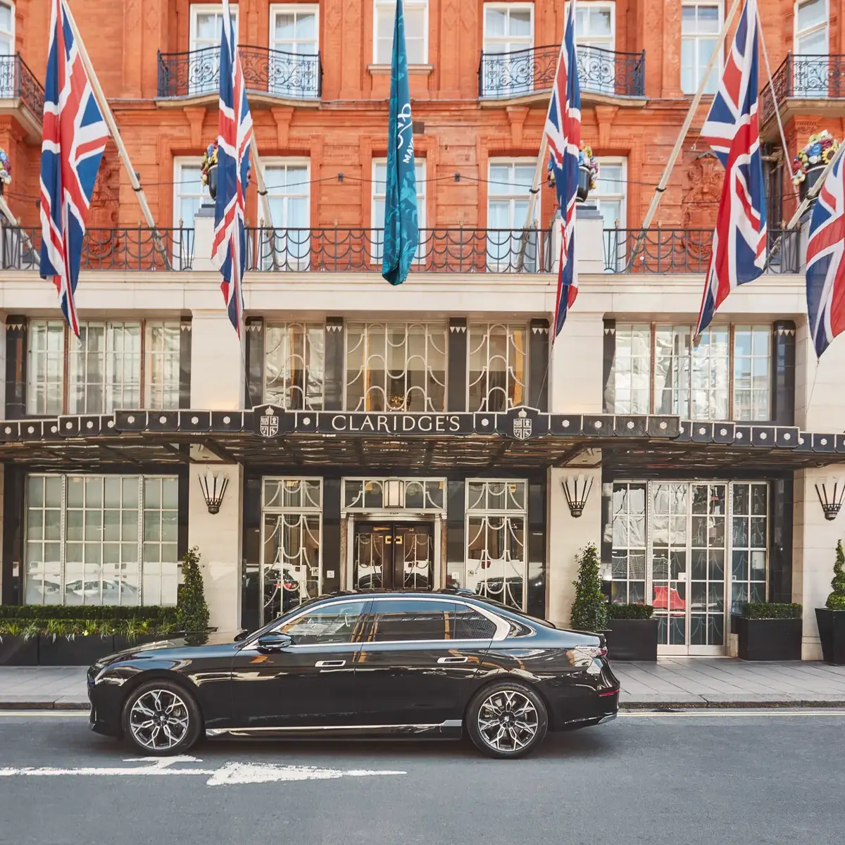 A sleek black luxury car parked outside the entrance of Claridge’s hotel in London, with Union Jack and teal flags hanging above the grand Art Deco façade of red brick and cream stone.