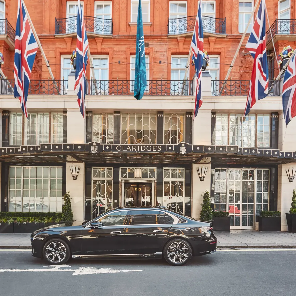A sleek black luxury car parked outside the entrance of Claridge’s hotel in London, with Union Jack and teal flags hanging above the grand Art Deco façade of red brick and cream stone.