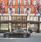 A sleek black luxury car parked outside the entrance of Claridge’s hotel in London, with Union Jack and teal flags hanging above the grand Art Deco façade of red brick and cream stone.