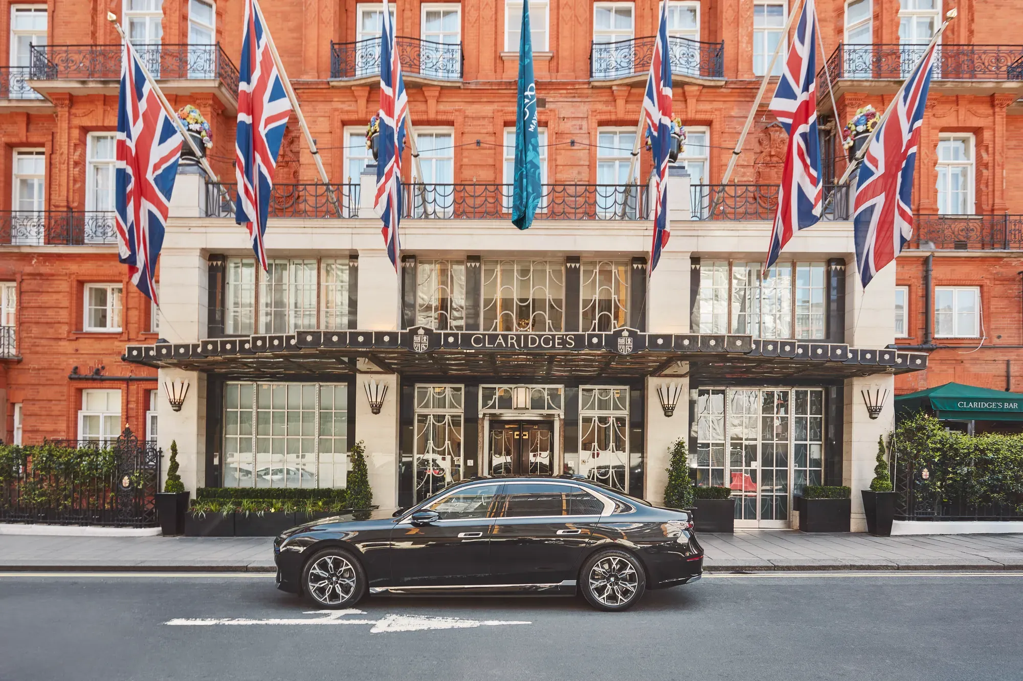A sleek black luxury car parked outside the entrance of Claridge’s hotel in London, with Union Jack and teal flags hanging above the grand Art Deco façade of red brick and cream stone.
