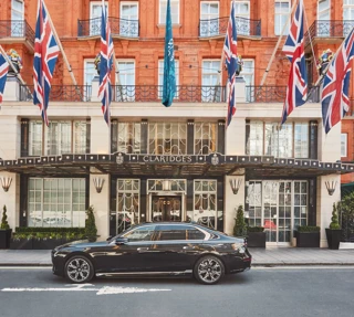 A sleek black luxury car parked outside the entrance of Claridge’s hotel in London, with Union Jack and teal flags hanging above the grand Art Deco façade of red brick and cream stone.