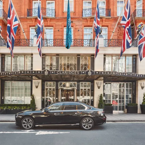 A sleek black luxury car parked outside the entrance of Claridge’s hotel in London, with Union Jack and teal flags hanging above the grand Art Deco façade of red brick and cream stone.