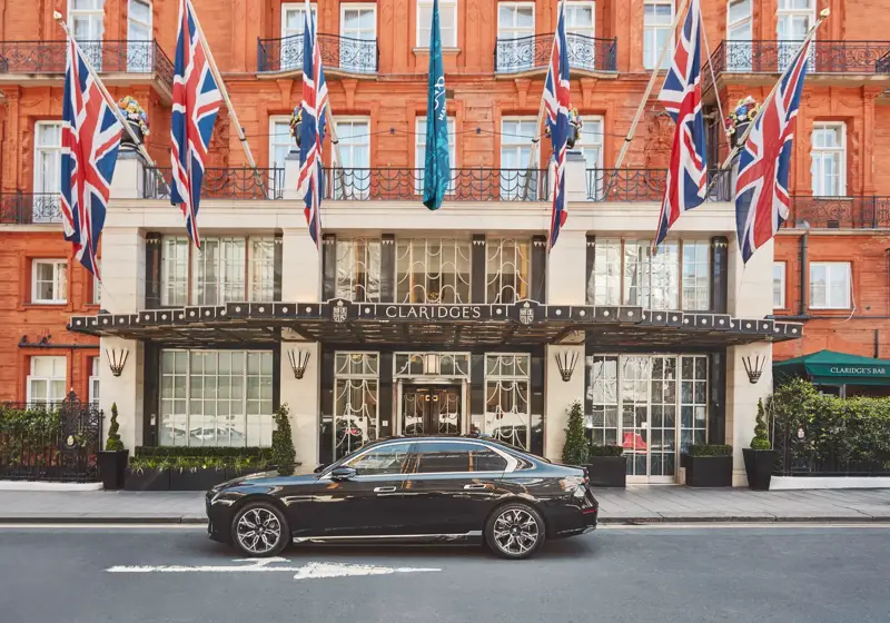 A sleek black luxury car parked outside the entrance of Claridge’s hotel in London, with Union Jack and teal flags hanging above the grand Art Deco façade of red brick and cream stone.