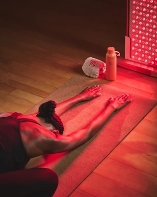 Guest stretches on yoga mat beside red light therapy panel, water bottle and towel nearby.
