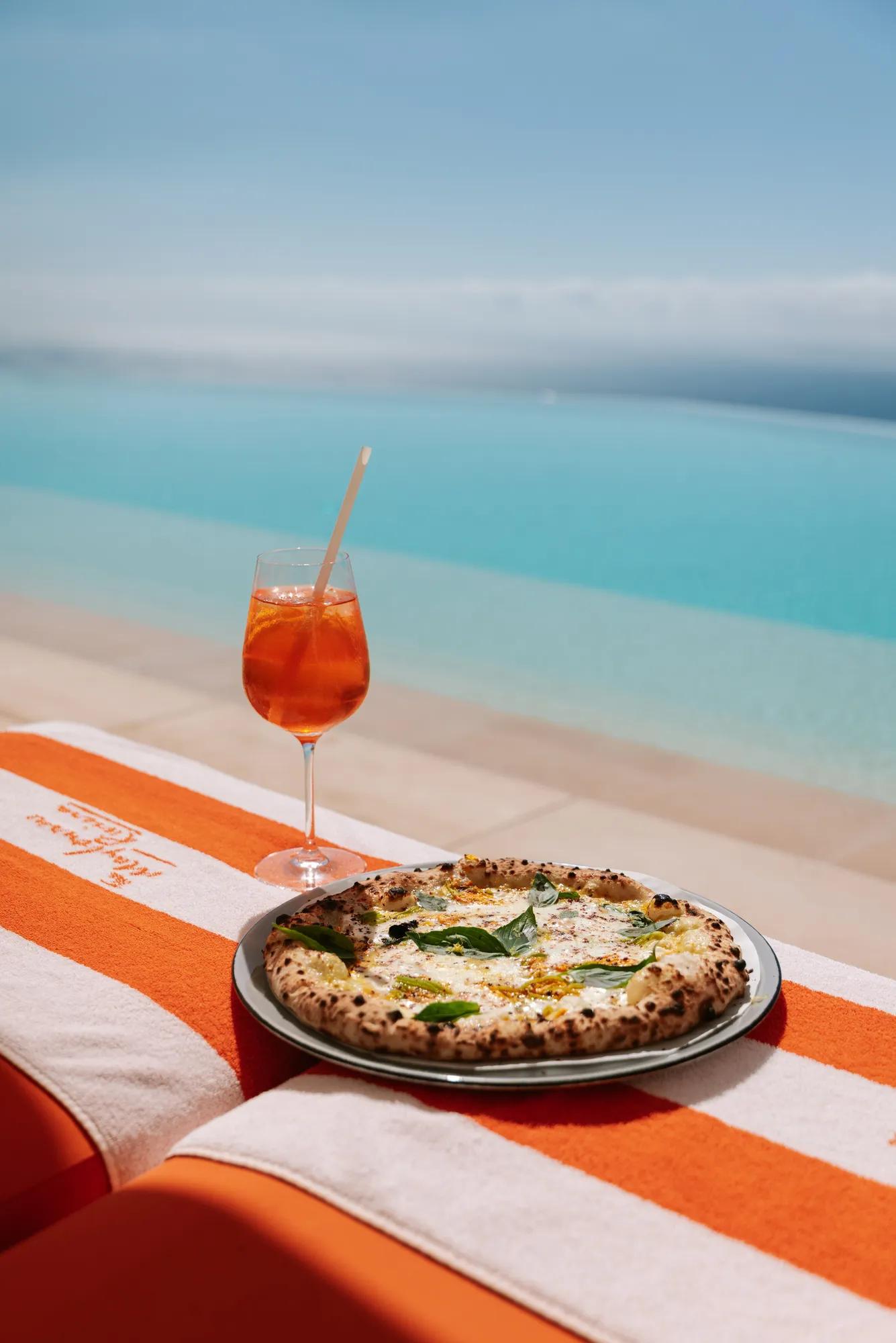 Un verre de cocktail orange et une pizza fraîchement cuite posés sur une serviette rayée orange et blanche, au bord d’une piscine à débordement avec vue sur la mer.