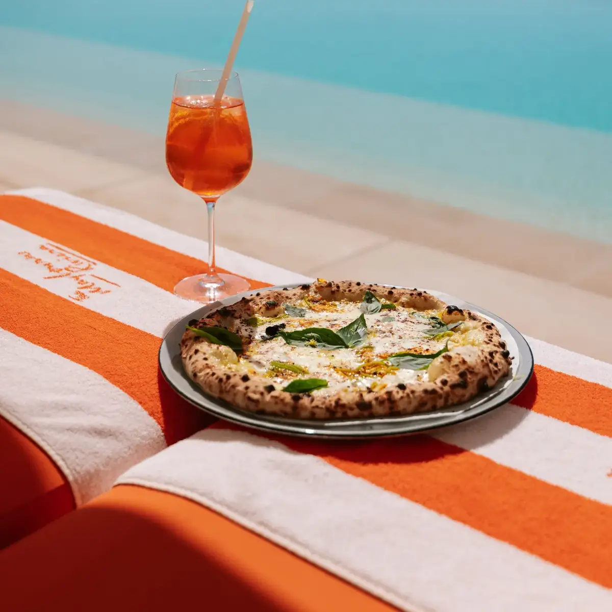 Un verre de cocktail orange et une pizza fraîchement cuite posés sur une serviette rayée orange et blanche, au bord d’une piscine à débordement avec vue sur la mer.