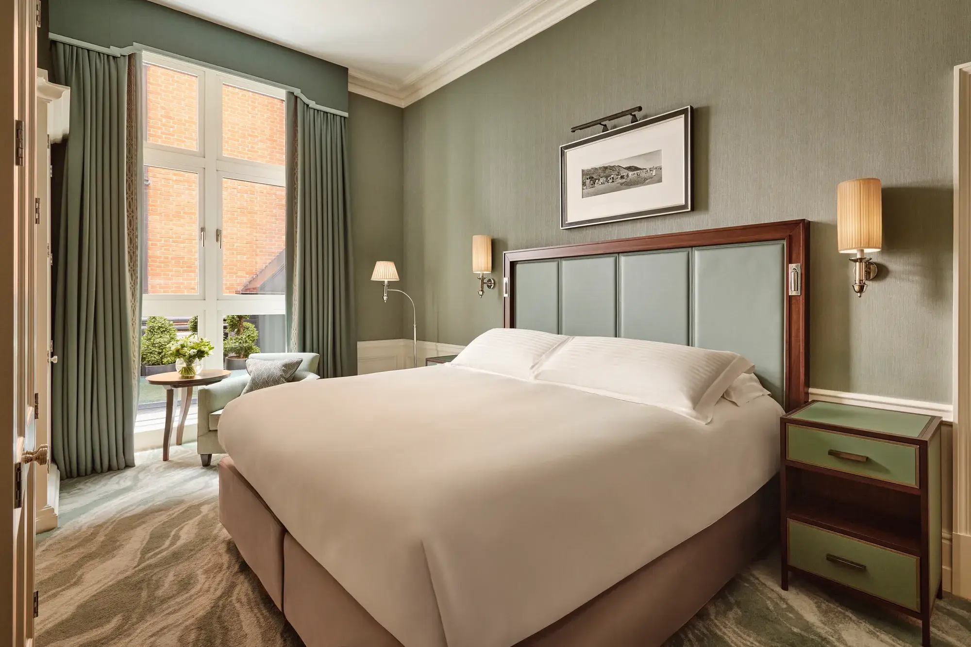 Soft green bedroom with a large bed, tall window with drapes, and a small seating area beside a round table.