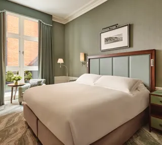 Soft green bedroom with a large bed, tall window with drapes, and a small seating area beside a round table.