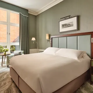 Soft green bedroom with a large bed, tall window with drapes, and a small seating area beside a round table.