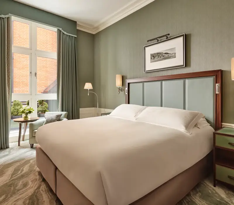 Soft green bedroom with a large bed, tall window with drapes, and a small seating area beside a round table.