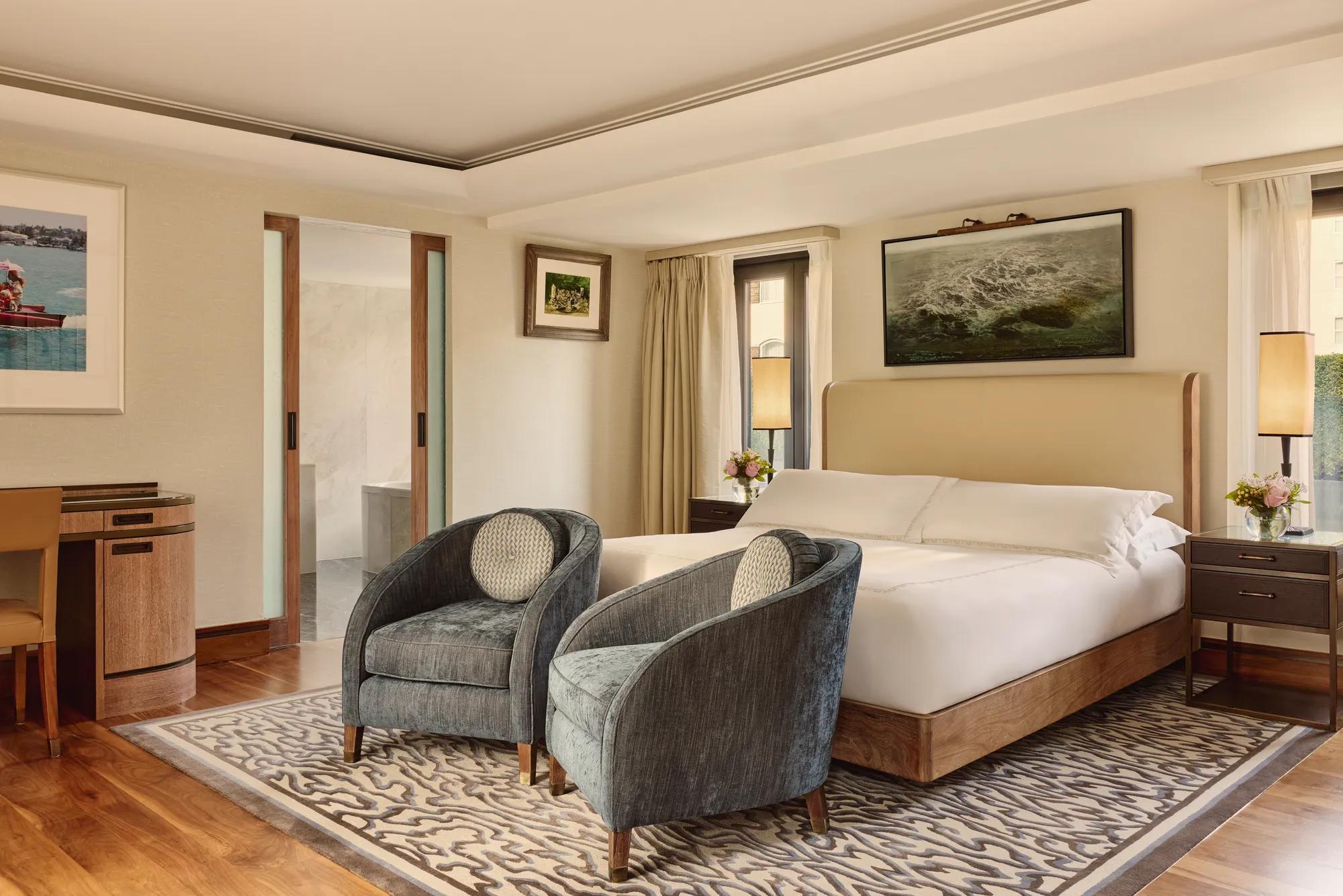 Spacious bedroom with upholstered bed, two blue armchairs, patterned rug, and sliding door to bathroom.