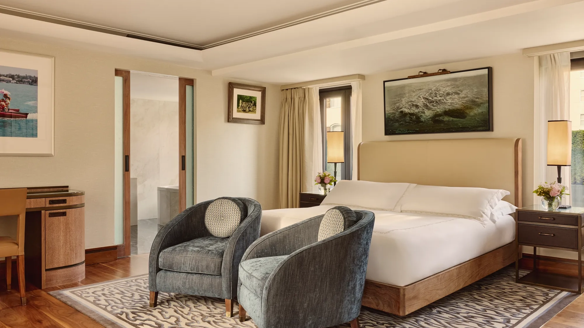 Spacious bedroom with upholstered bed, two blue armchairs, patterned rug, and sliding door to bathroom.