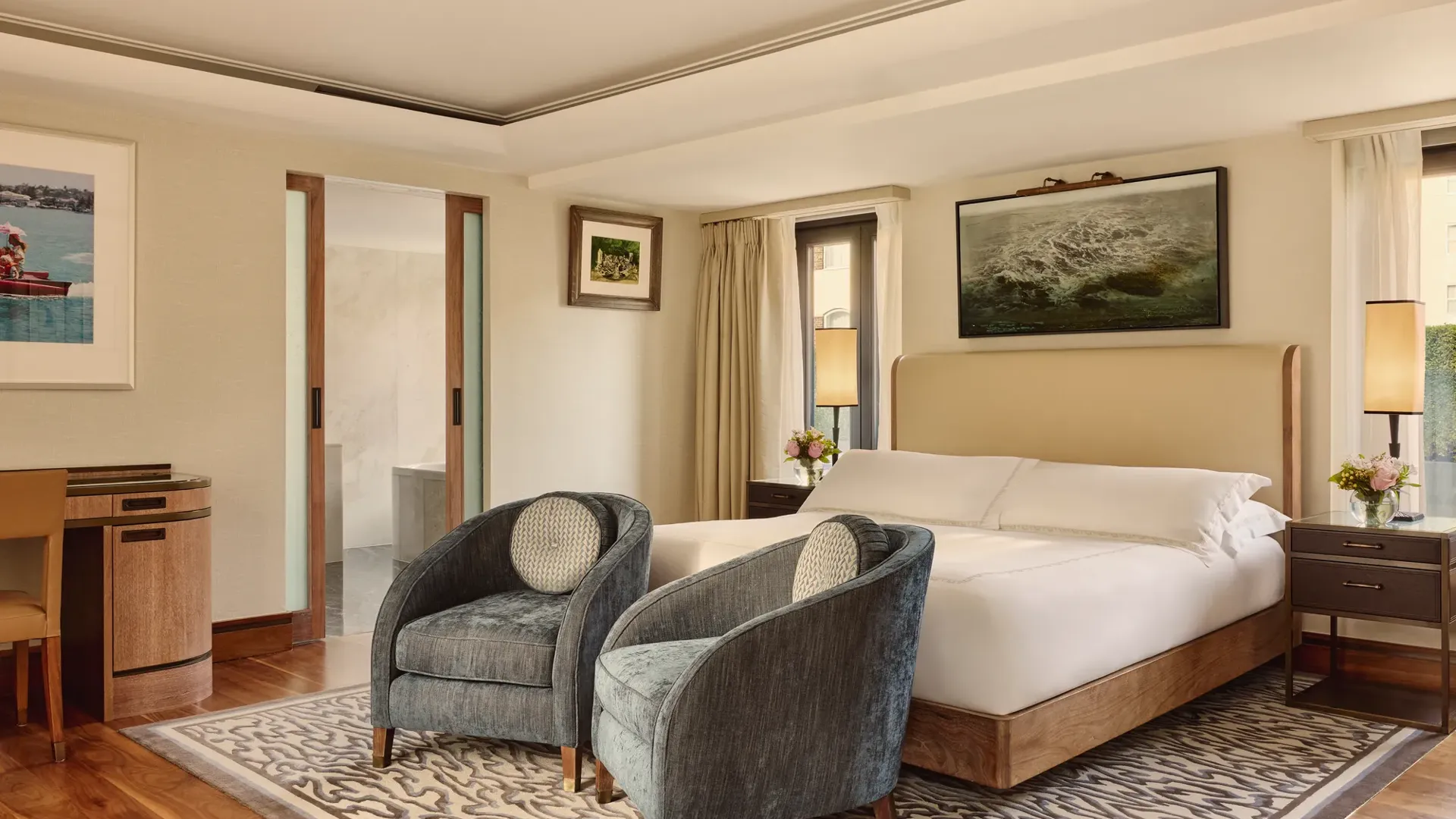 Spacious bedroom with upholstered bed, two blue armchairs, patterned rug, and sliding door to bathroom.