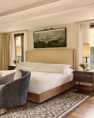 Spacious bedroom with upholstered bed, two blue armchairs, patterned rug, and sliding door to bathroom.