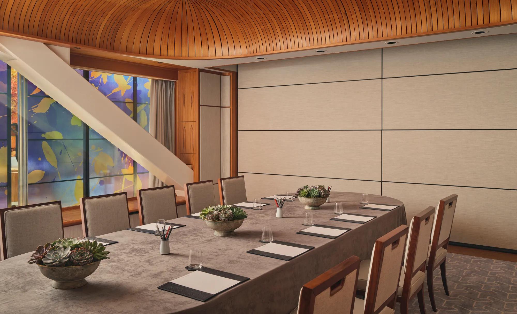 Boardroom setup with long table, notepads, succulents, and patterned glass window under curved wood ceiling.