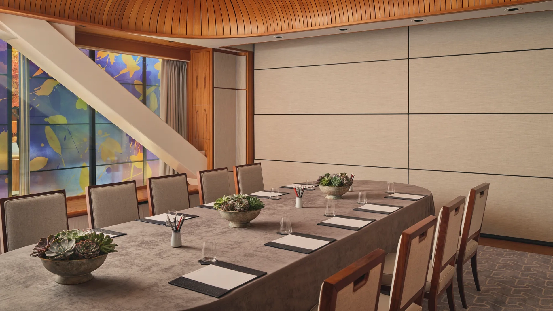 Boardroom setup with long table, notepads, succulents, and patterned glass window under curved wood ceiling.
