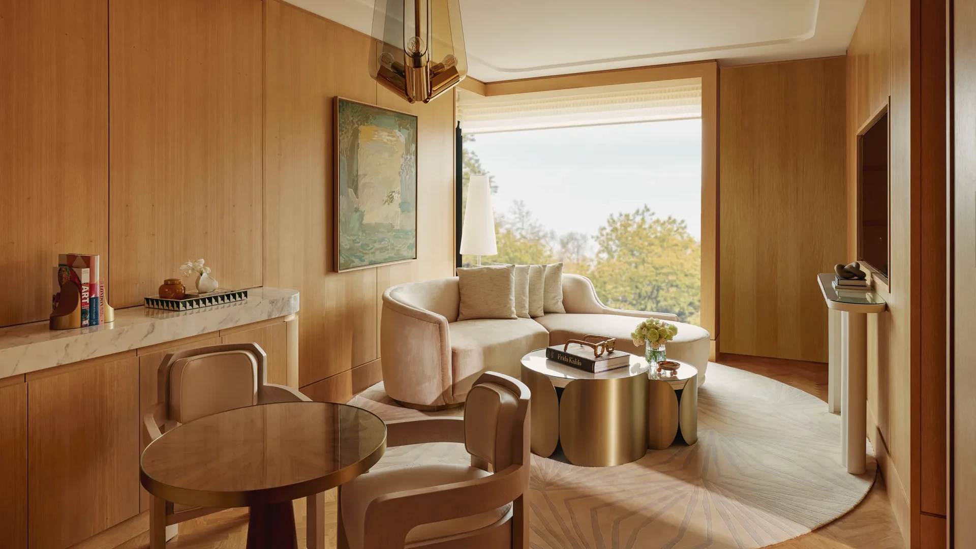 Elegant living area with curved sofa, round dining table, warm wood panelling, sculptural lighting and large window with leafy views.