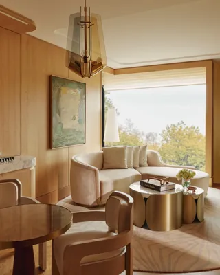 Elegant living area with curved sofa, round dining table, warm wood panelling, sculptural lighting and large window with leafy views.