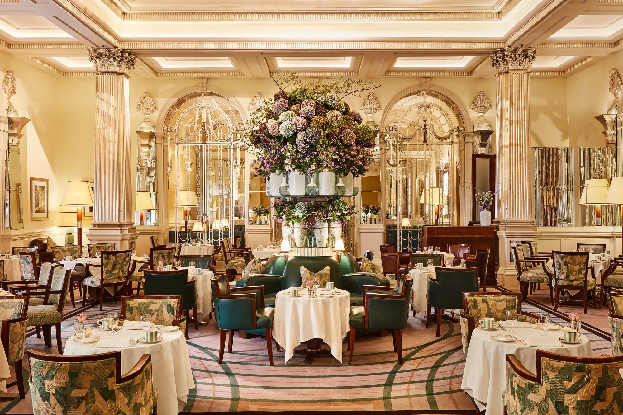 Elegant dining room at Claridge’s with round tables, green leather chairs, mirrored walls, and a large floral arrangement at the centre.