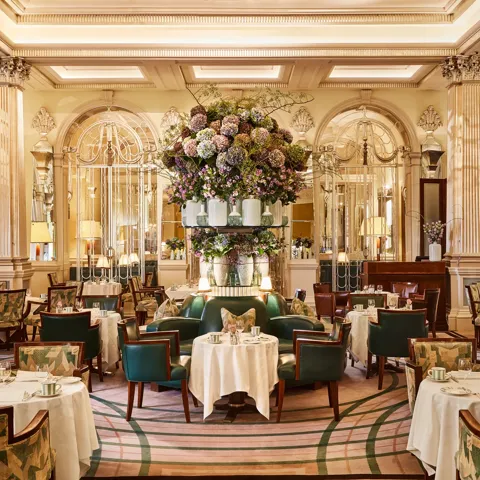 Elegant dining room at Claridge’s with round tables, green leather chairs, mirrored walls, and a large floral arrangement at the centre.