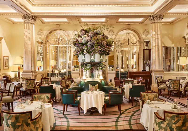 Elegant dining room at Claridge’s with round tables, green leather chairs, mirrored walls, and a large floral arrangement at the centre.