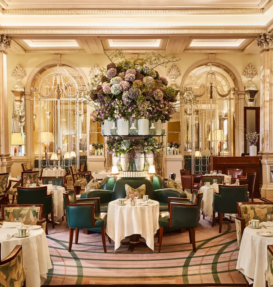 Elegant dining room at Claridge’s with round tables, green leather chairs, mirrored walls, and a large floral arrangement at the centre.