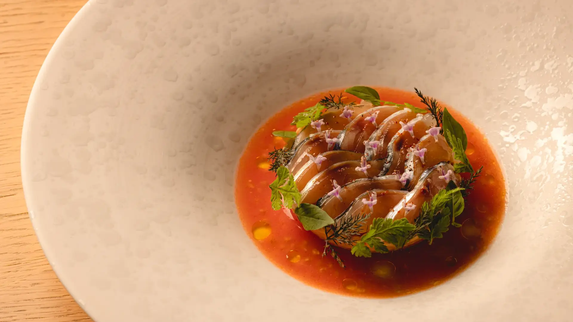 Plated dish of sliced fish in a vibrant tomato broth, garnished with herbs and edible flowers in a wide white bowl.