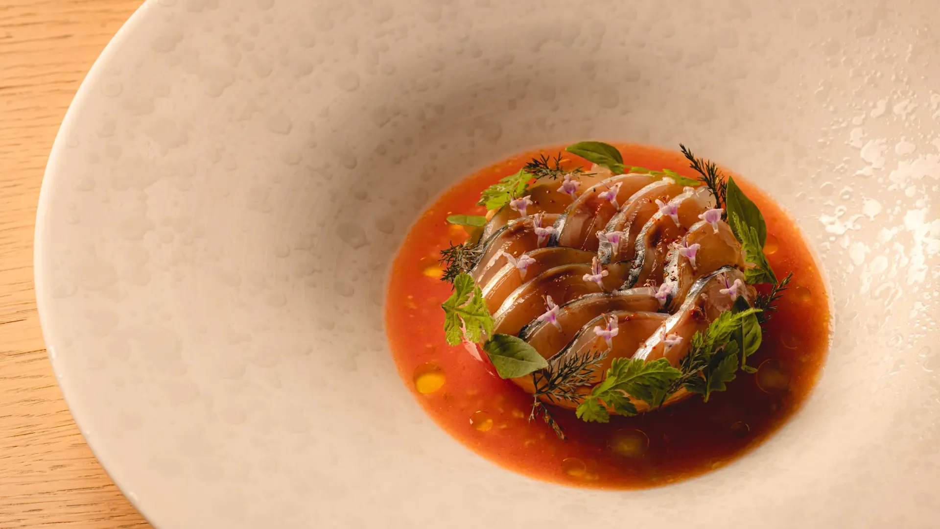 Plated dish of sliced fish in a vibrant tomato broth, garnished with herbs and edible flowers in a wide white bowl.