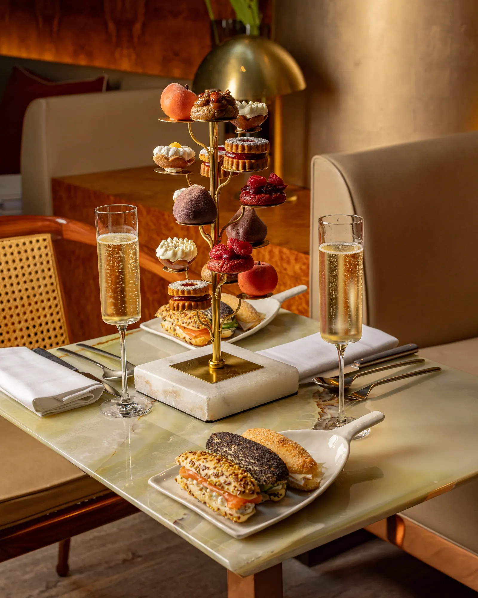 Afternoon tea table with champagne, sandwiches, and a gold tiered stand of miniature pastries in a softly lit setting.