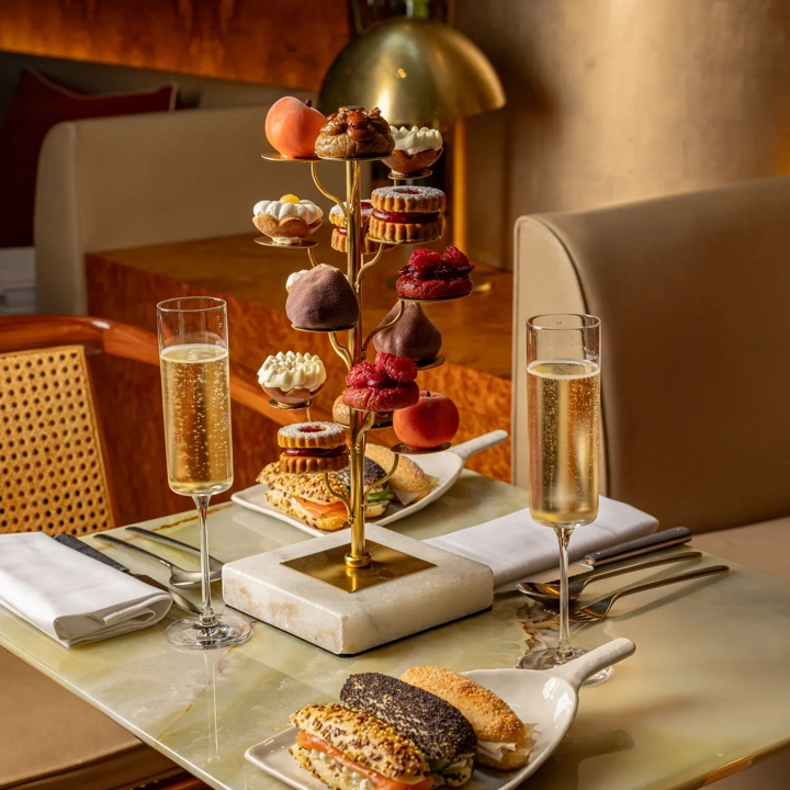 Afternoon tea table with champagne, sandwiches, and a gold tiered stand of miniature pastries in a softly lit setting.
