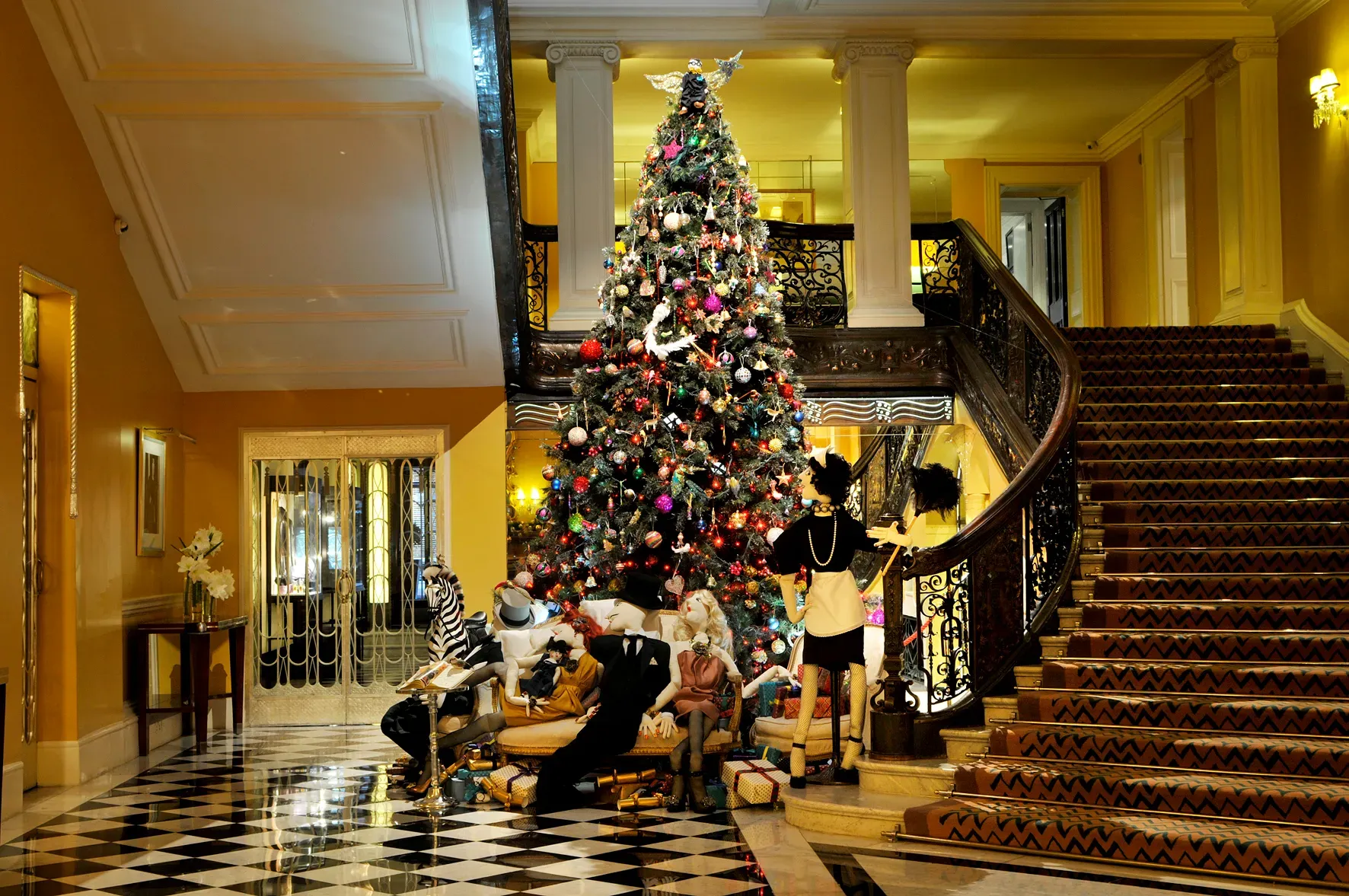 Grand hotel lobby with sweeping staircase and checkered floor, featuring large decorated Christmas tree surrounded by mannequins and gifts.