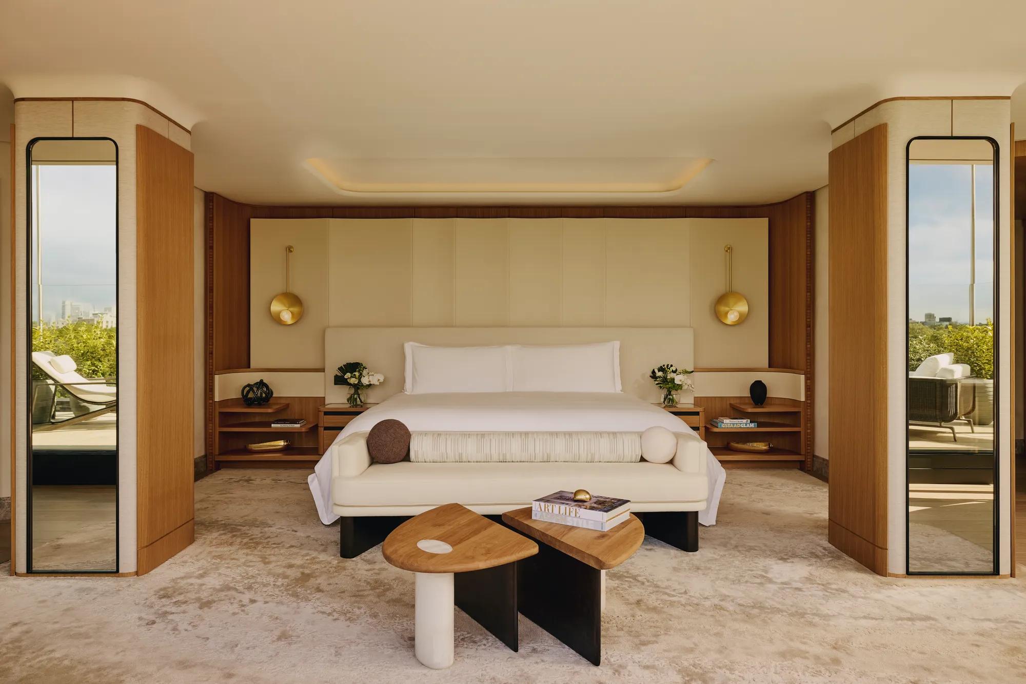 Spacious bedroom with a large bed, cream headboard, modern gold pendant lights, mirrored columns, and geometric wooden tables, opening onto a terrace with greenery outside.
