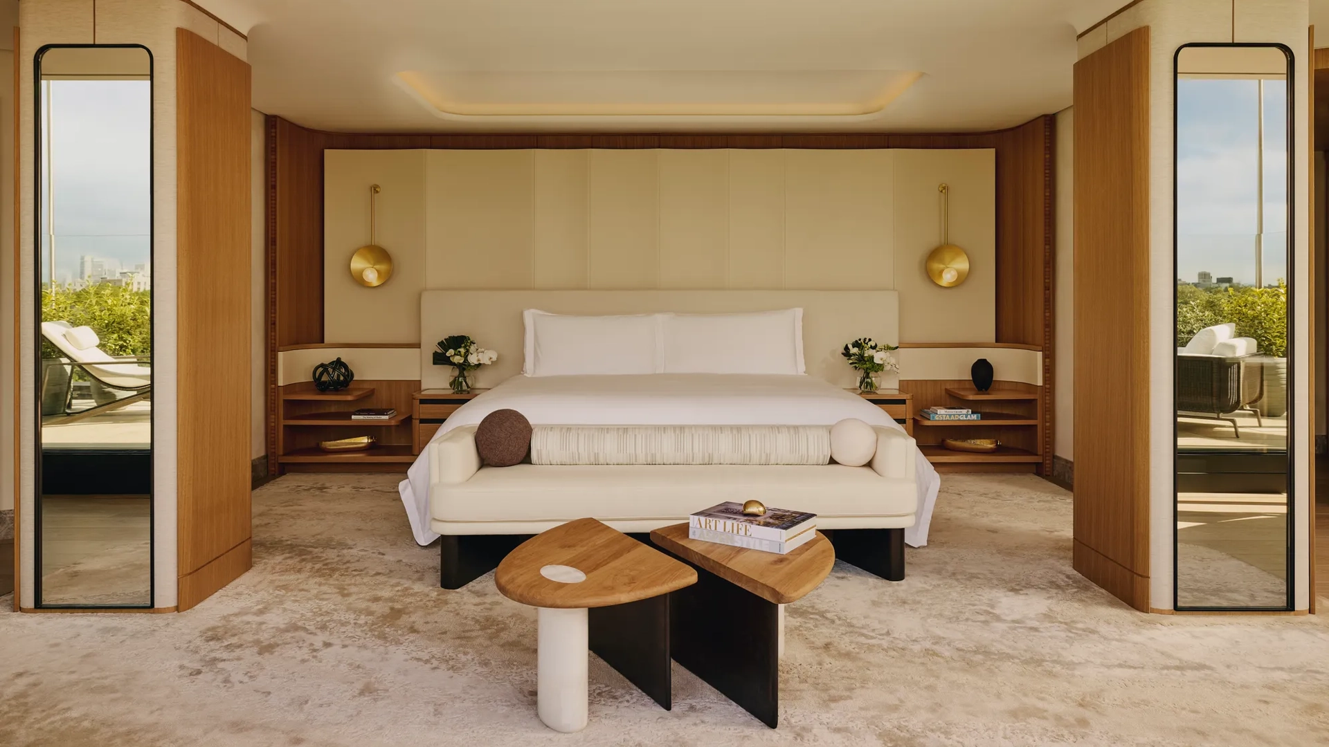 Spacious bedroom with a large bed, cream headboard, modern gold pendant lights, mirrored columns, and geometric wooden tables, opening onto a terrace with greenery outside.