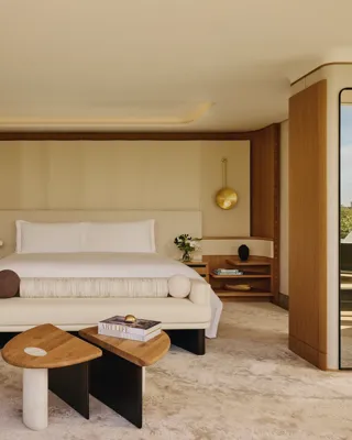 Spacious bedroom with a large bed, cream headboard, modern gold pendant lights, mirrored columns, and geometric wooden tables, opening onto a terrace with greenery outside.